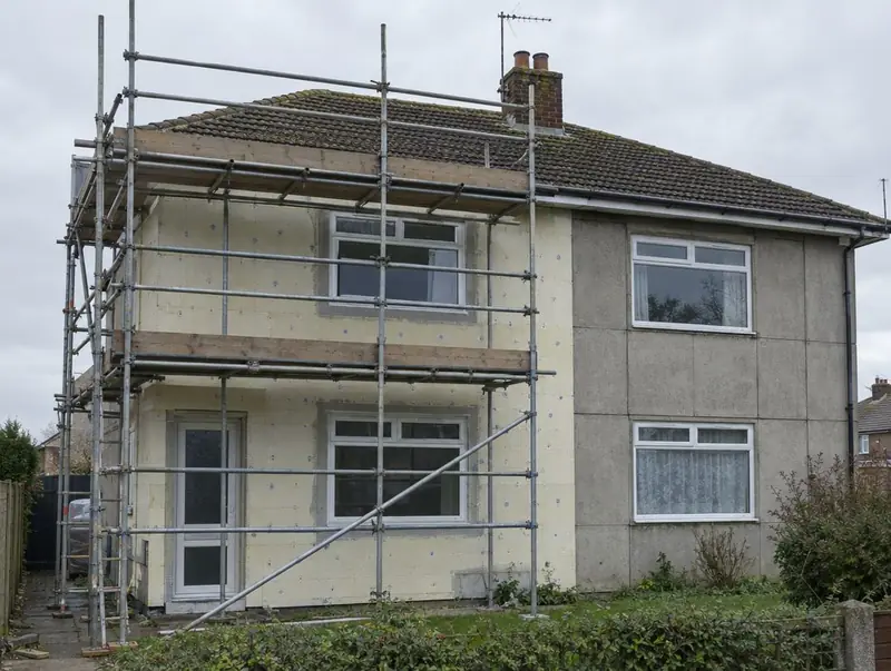 Post-war concrete semi-detached house half clad in insulation and mesh under scaffolding, the other half showing panel joints